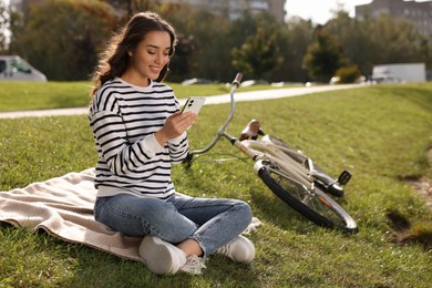 Photo of Young woman sitting on green grass and using smartphone near bicycle in park, space for text
