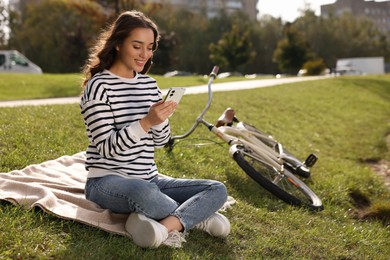 Young woman sitting on green grass and using smartphone near bicycle in park, space for text