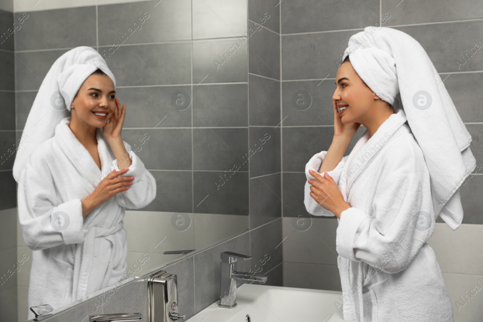 Photo of Beautiful young woman in bathrobe with towel on head near mirror at home