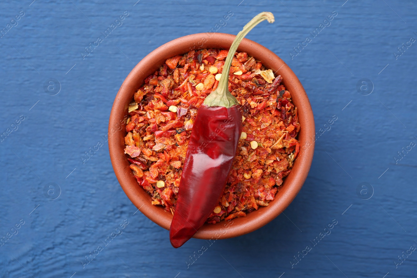 Photo of Chili pepper flakes in bowl and pod on blue wooden table, top view