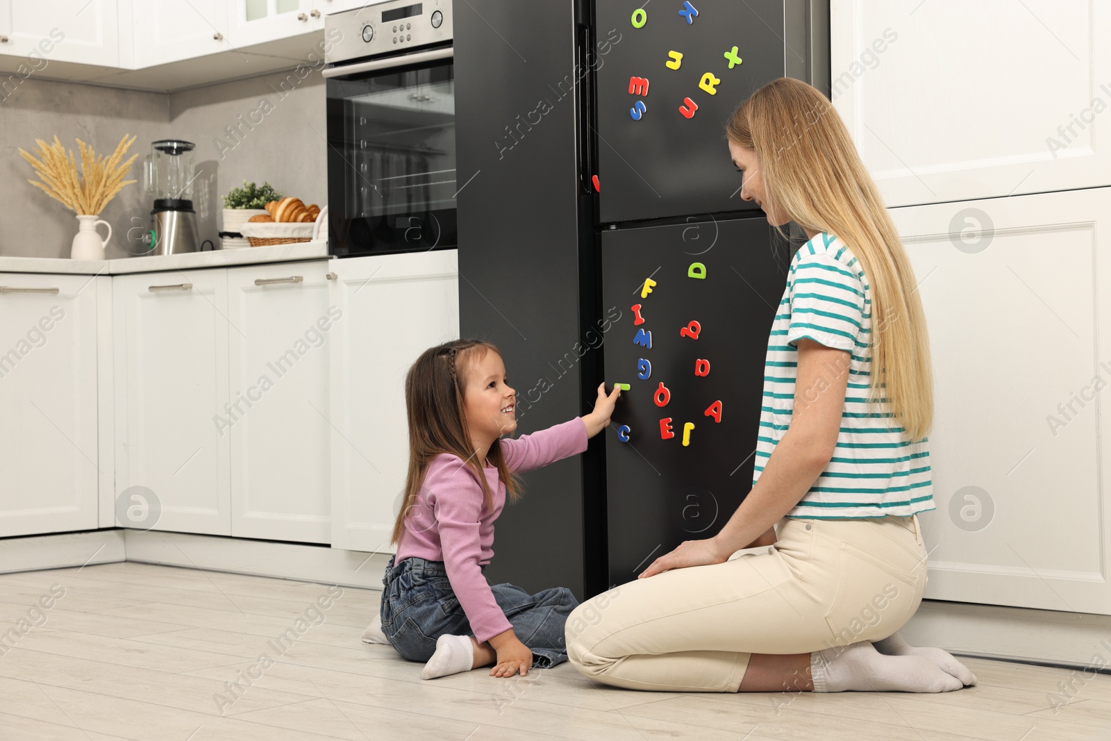 Photo of Mom and daughter putting magnetic letters on fridge at home. Learning alphabet