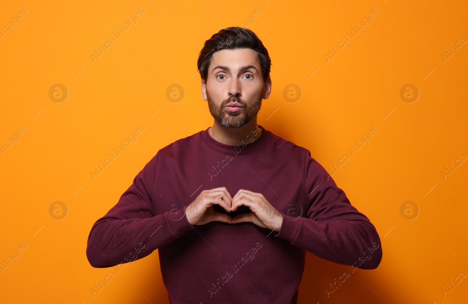 Photo of Handsome man making heart with hands and blowing kiss on orange background