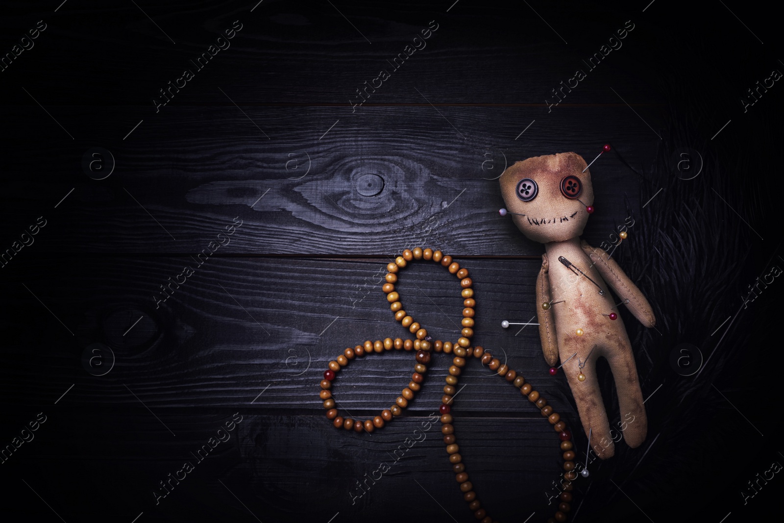 Image of Voodoo doll pierced with pins, beads and feather on black wooden table, flat lay