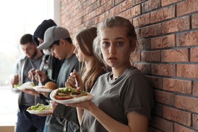 Photo of Young poor woman and other people with food at brick wall indoors