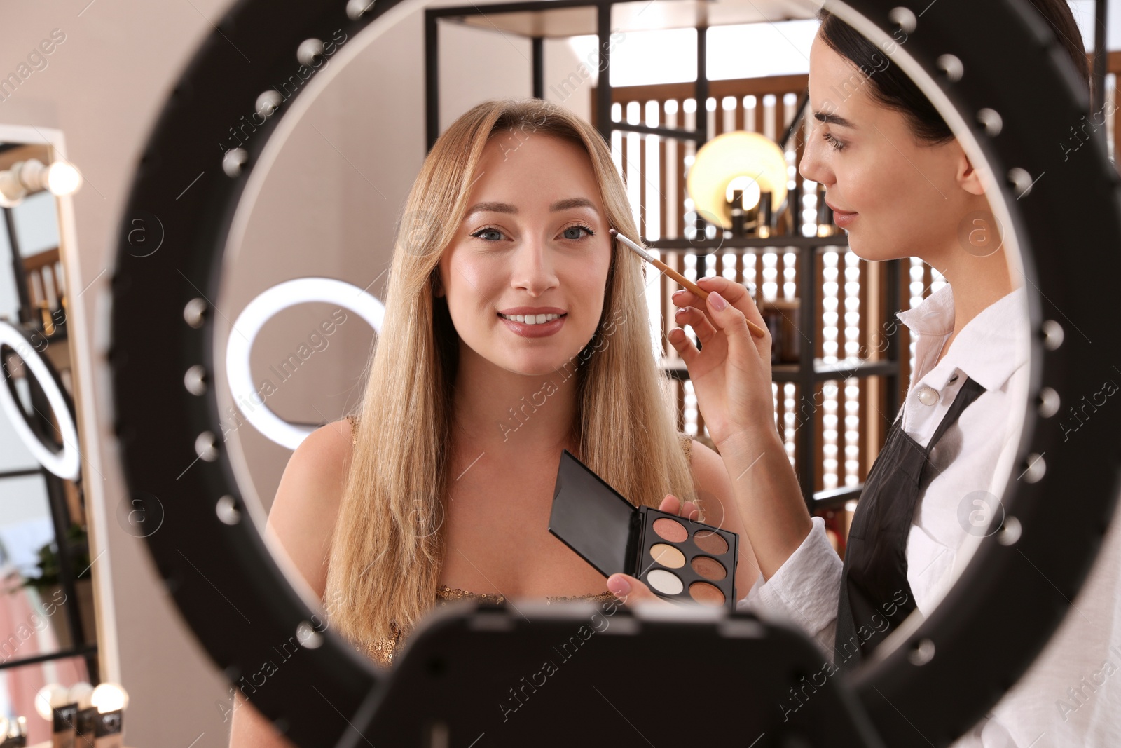 Photo of Professional makeup artist working with beautiful young woman  in salon, view through ring lamp