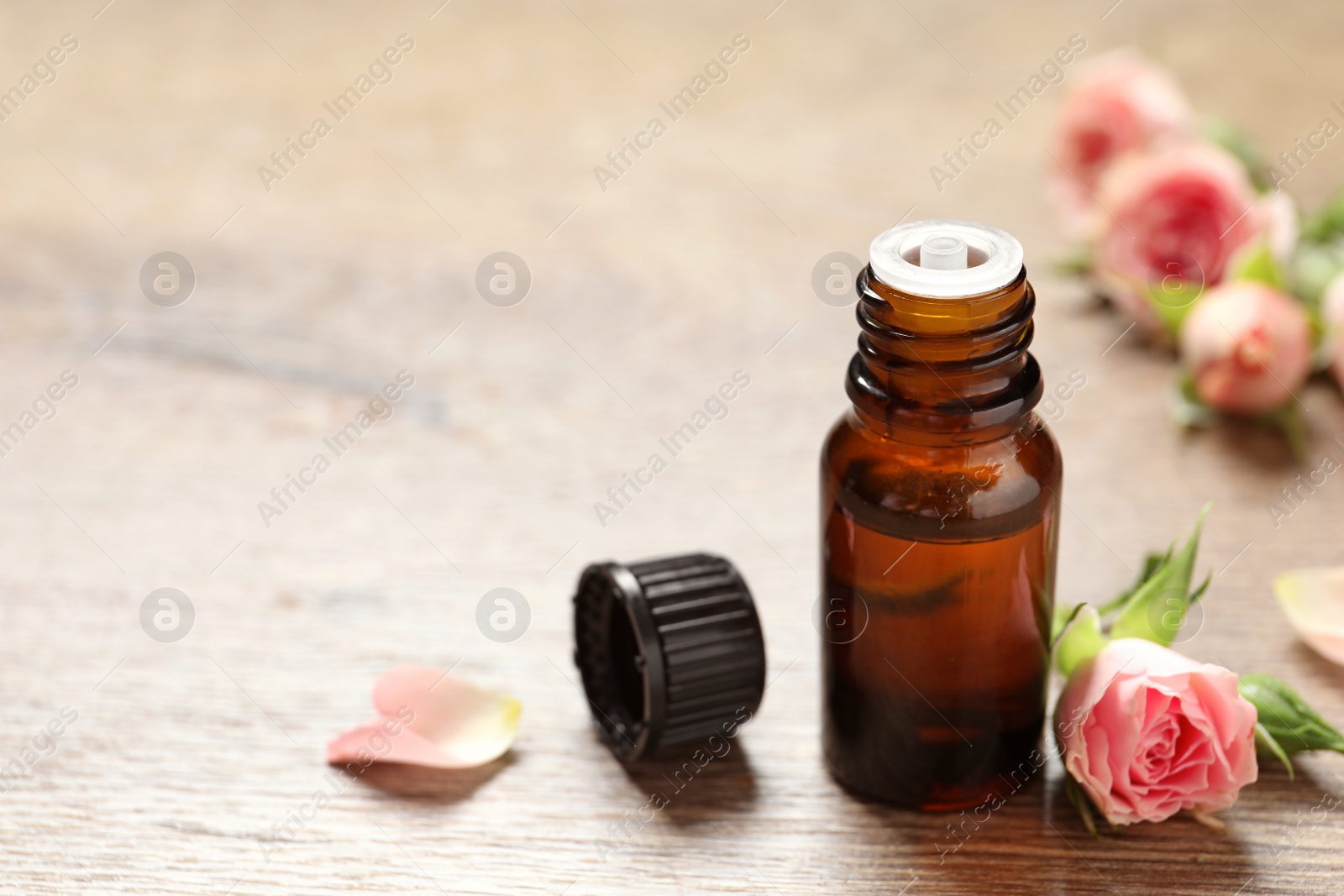 Photo of Bottle of rose essential oil and fresh flowers on wooden table, space for text