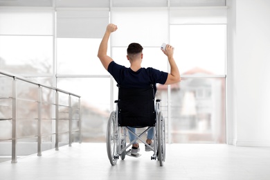 Emotional young man in wheelchair with mobile phone against window indoors