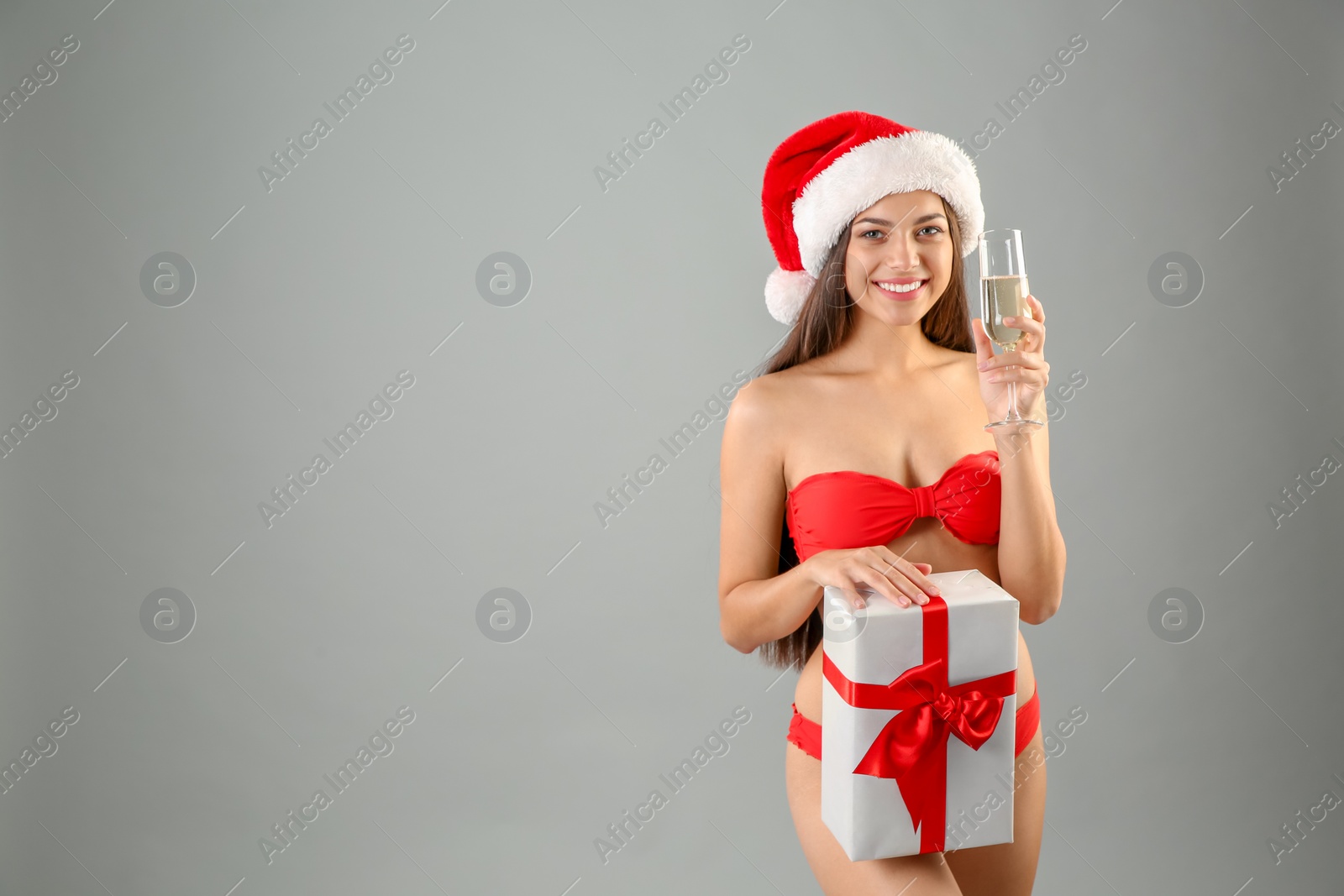 Photo of Young woman in Santa hat and swimsuit holding gift box with glass of champagne on grey background. Christmas celebration