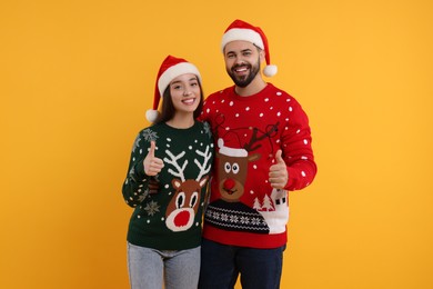 Photo of Happy young couple in Christmas sweaters and Santa hats showing thumbs up on orange background