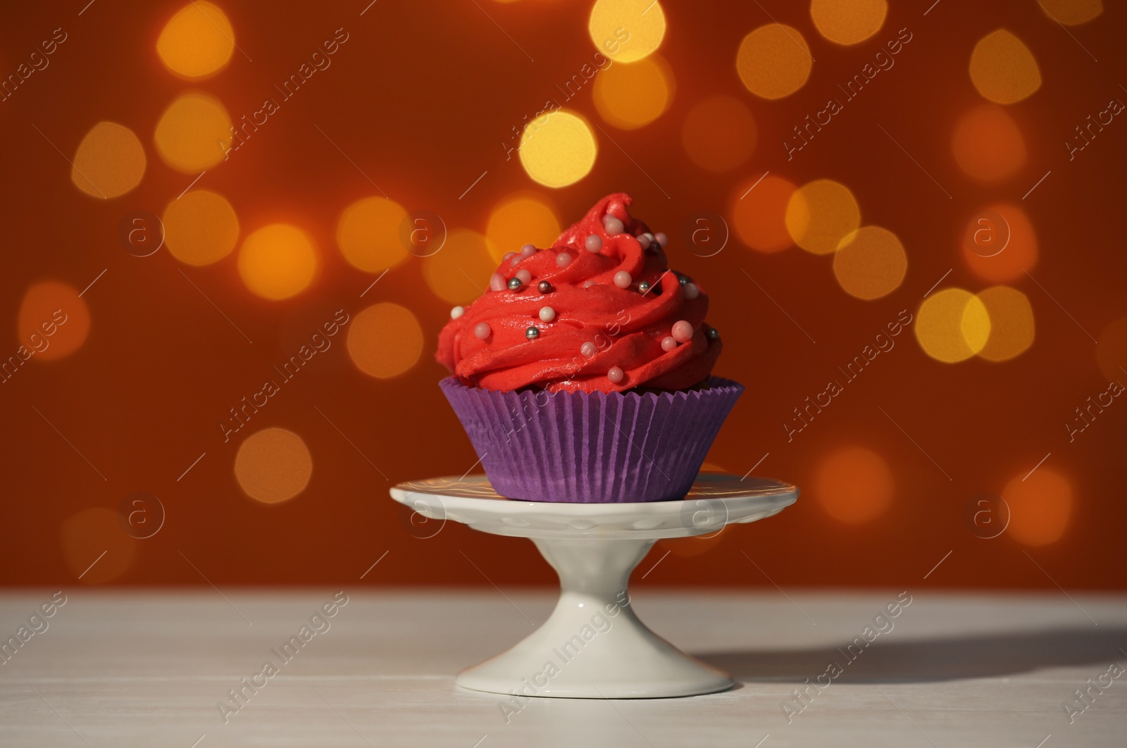 Photo of Stand with tasty cupcake on white table against blurred lights