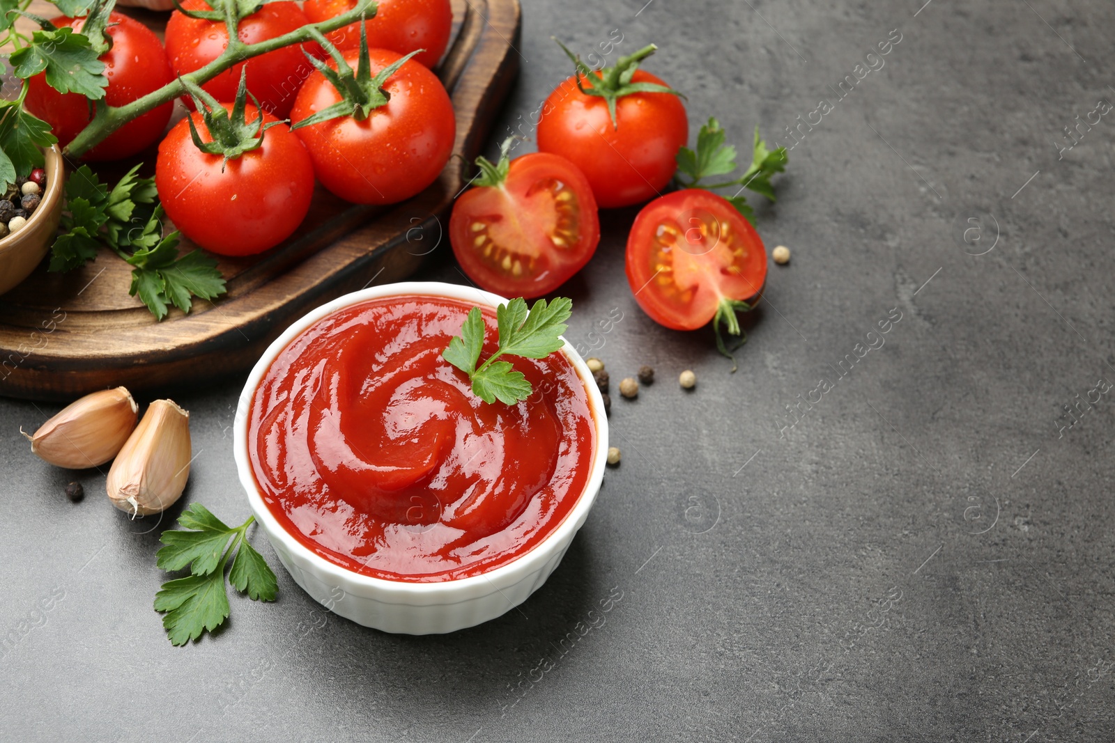 Photo of Delicious ketchup in bowl, tomatoes, parsley and garlic on grey table, closeup. Space for text