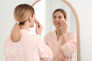 Beautiful woman removing makeup with cotton pad near mirror indoors