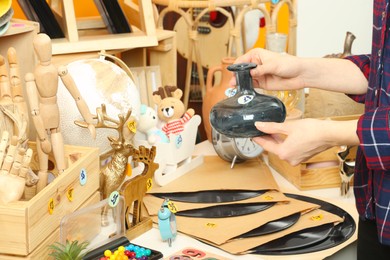 Photo of Woman holding vase near table with different stuff indoors, closeup. Garage sale