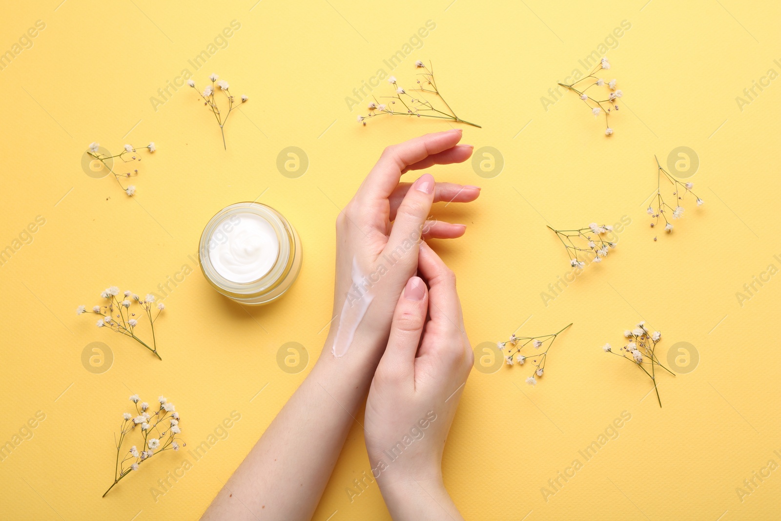 Photo of Woman applying hand cream and flowers on yellow background, top view