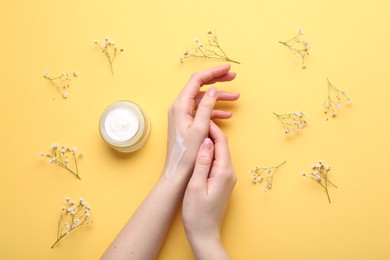 Woman applying hand cream and flowers on yellow background, top view