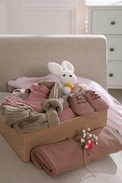 Photo of Wooden crate with children's clothes, shoes, toy bunny and pacifier on sofa in room