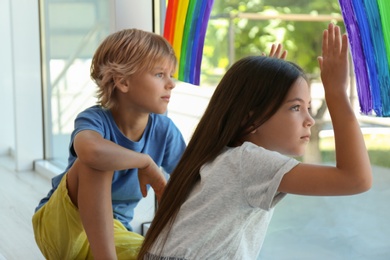 Photo of Little children sitting near rainbow painting on window indoors. Stay at home concept