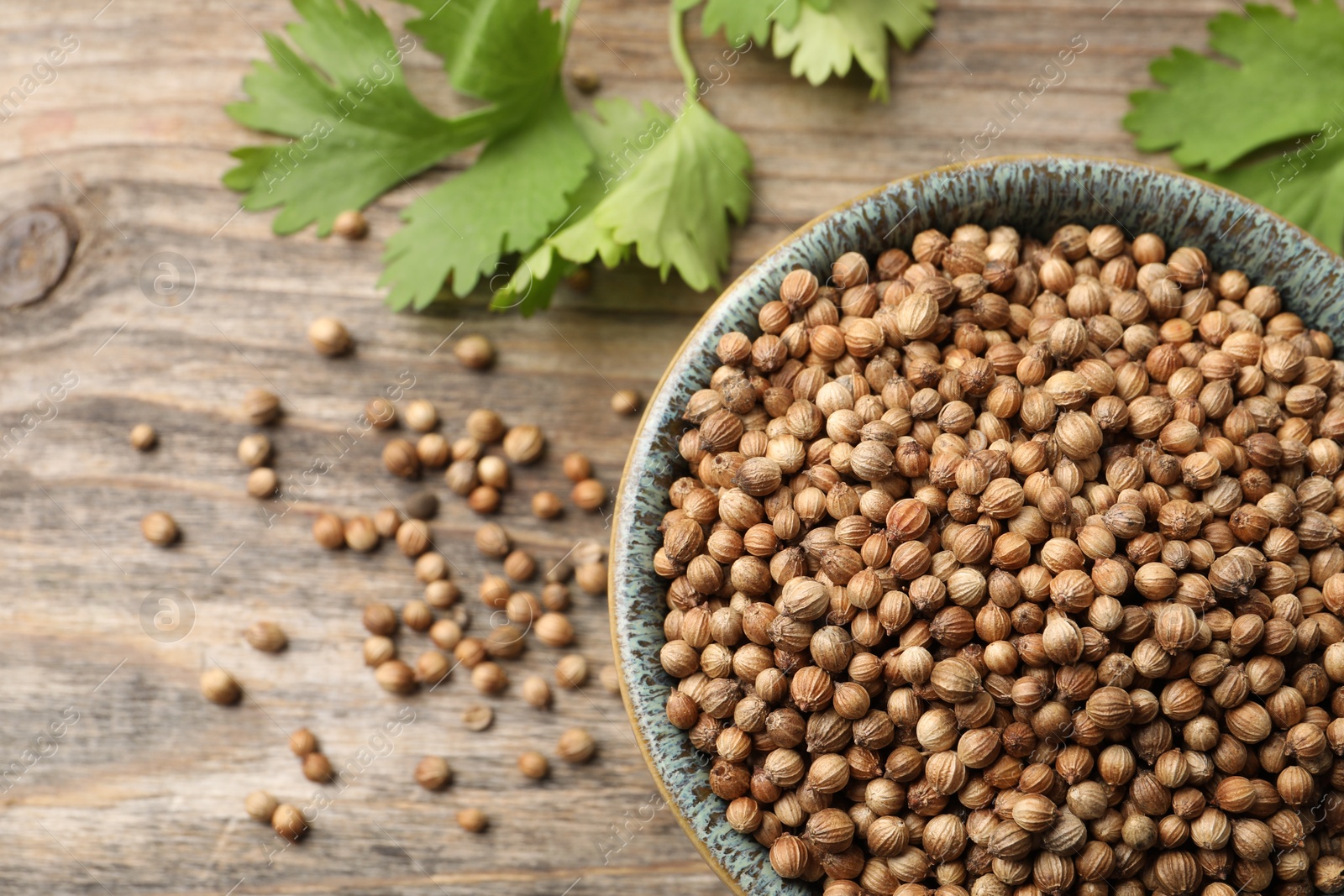 Photo of Dried coriander seeds in bowl and green leaves on wooden table, flat lay. Space for text