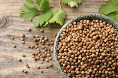 Photo of Dried coriander seeds in bowl and green leaves on wooden table, flat lay. Space for text