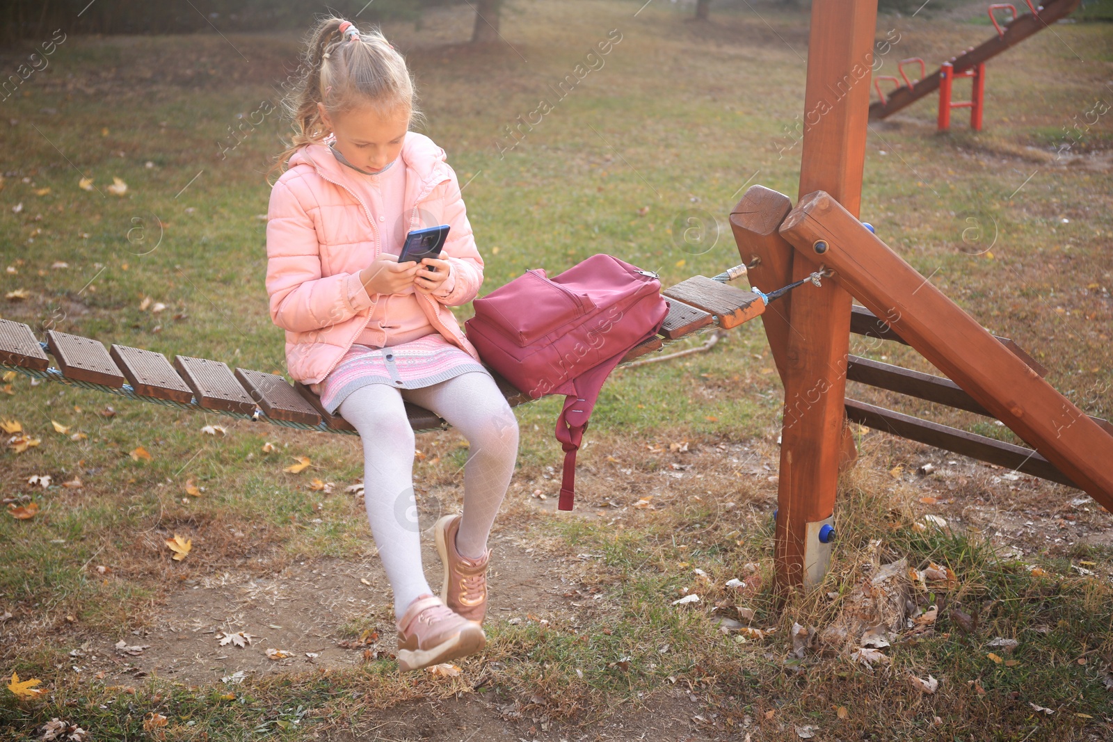 Photo of Cute little girl with backpack and smartphone on wooden swing outdoors