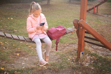 Photo of Cute little girl with backpack and smartphone on wooden swing outdoors