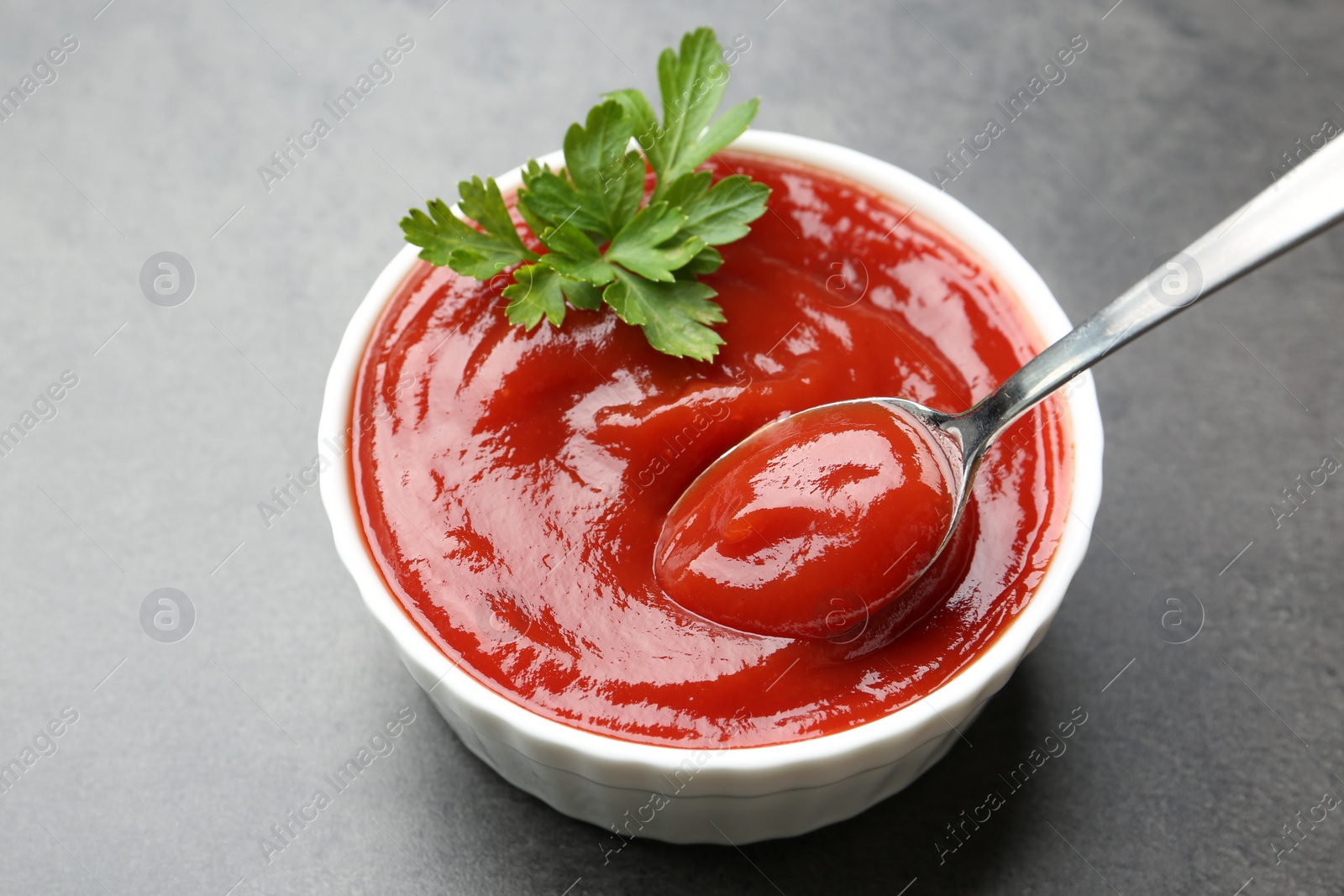 Photo of Taking delicious tomato ketchup with spoon from bowl at grey textured table, closeup