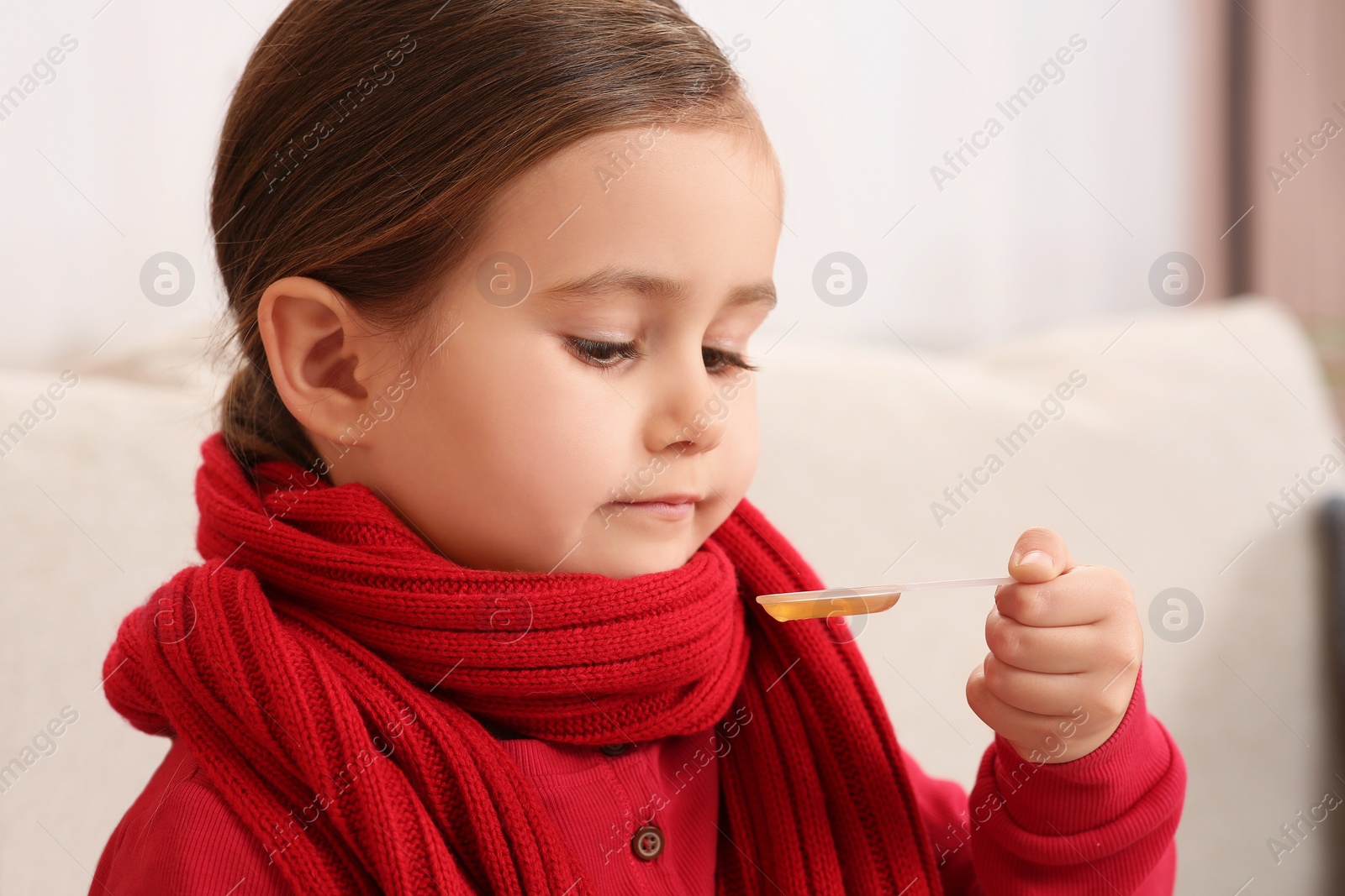 Photo of Cute girl holding measuring spoon with cough syrup on sofa indoors. Effective medicine