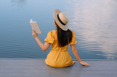 Photo of Woman reading book on pier near lake, back view