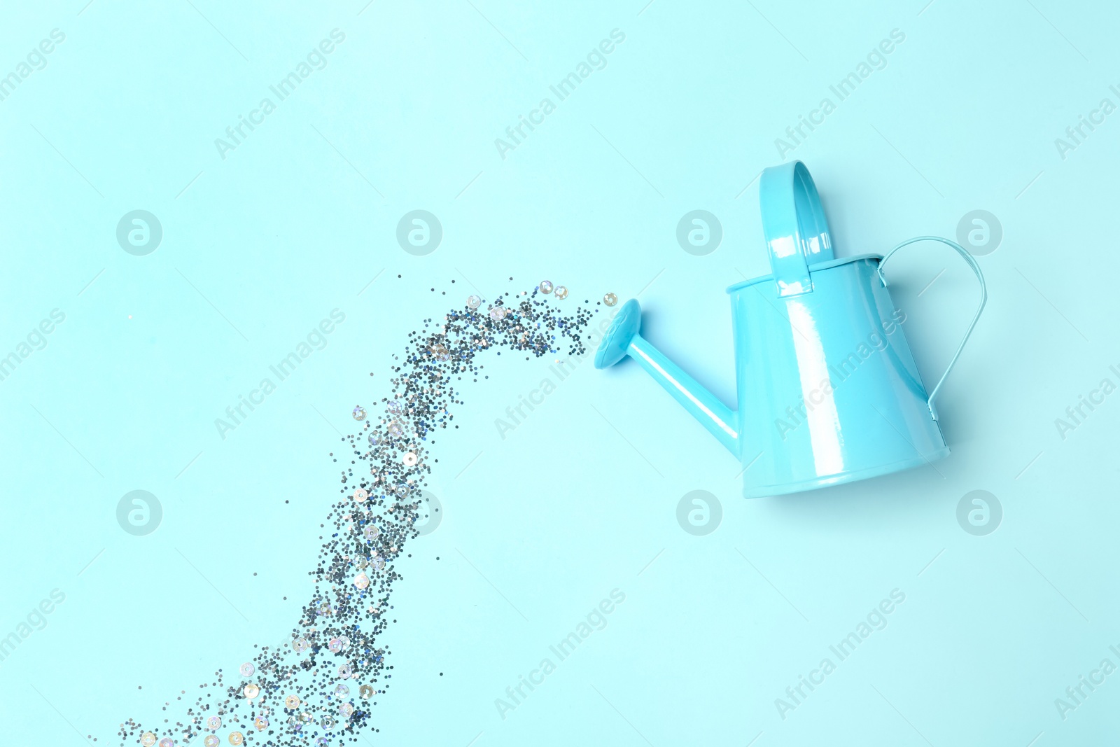 Photo of Watering can with silver glitter confetti imitating water  flow on cyan background, flat lay