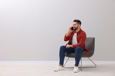 Handsome man talking on smartphone while sitting in armchair near light grey wall indoors, space for text