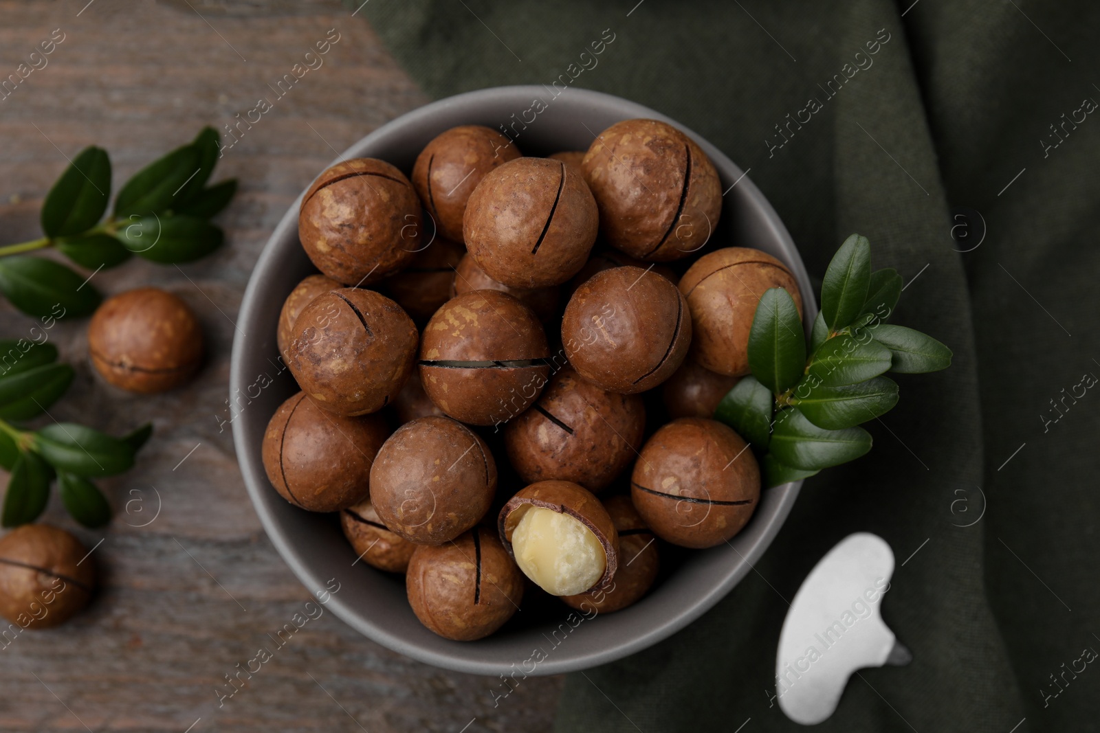 Photo of Tasty Macadamia nuts in bowl and green twigs on wooden table, flat lay