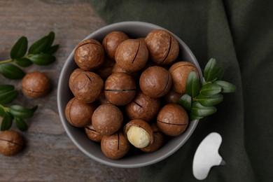 Tasty Macadamia nuts in bowl and green twigs on wooden table, flat lay