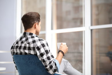 Man with cup of drink resting near window at home