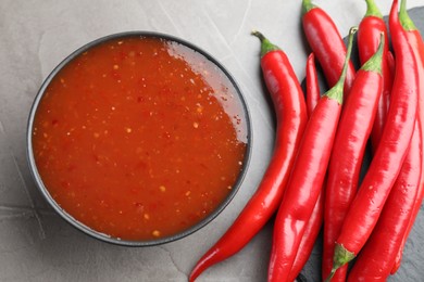 Photo of Spicy chili sauce in bowl and fresh peppers on grey table, flat lay