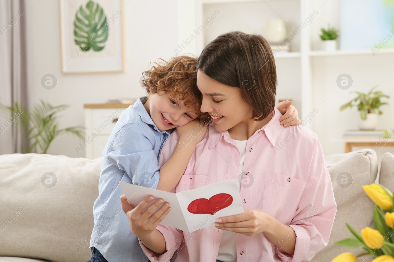 Photo of Little son congratulating his mom with Mother`s day at home. Woman holding handmade greeting card