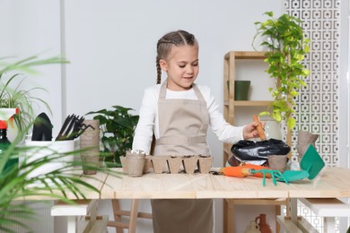 Photo of Little girl adding soil into peat pots at wooden table in room. Growing vegetable seeds