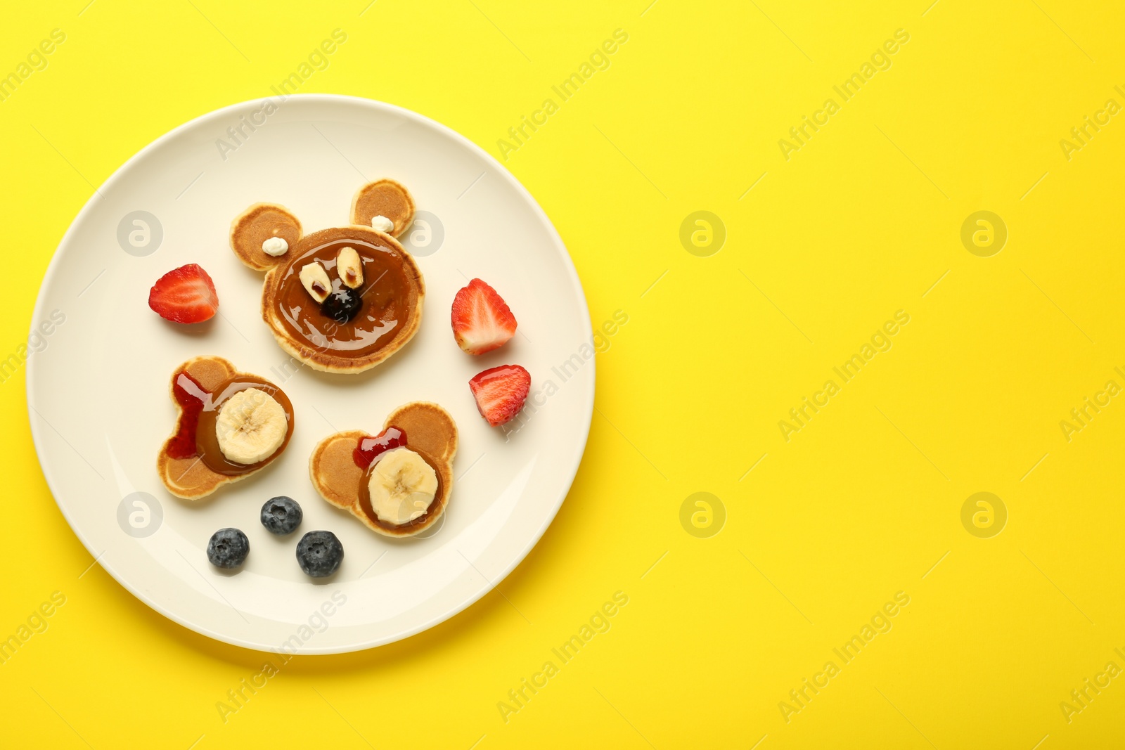 Photo of Creative serving for kids. Plate with cute bears made of pancakes, berries, banana and chocolate paste on yellow background, top view. Space for text