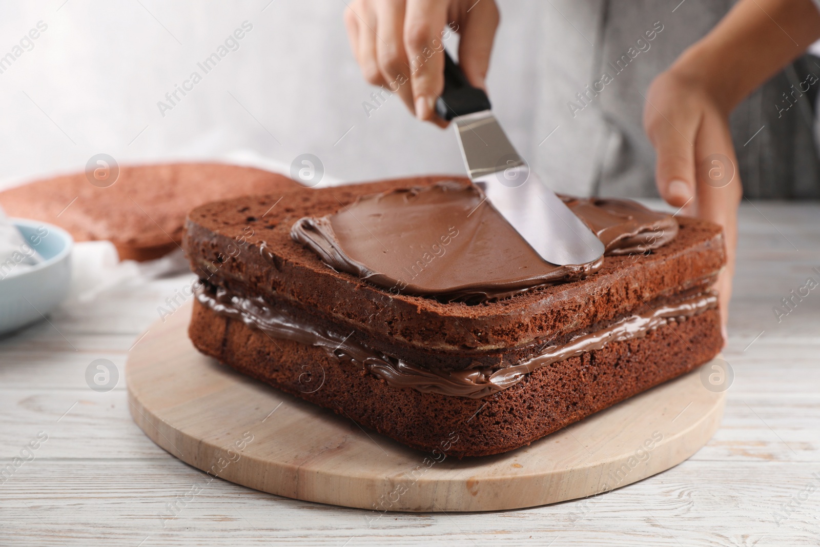 Photo of Woman smearing sponge cake with chocolate cream at white wooden table, closeup