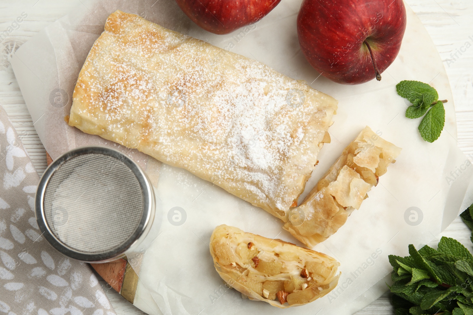 Photo of Delicious strudel, powdered sugar, mint and fresh apples on white wooden table, flat lay