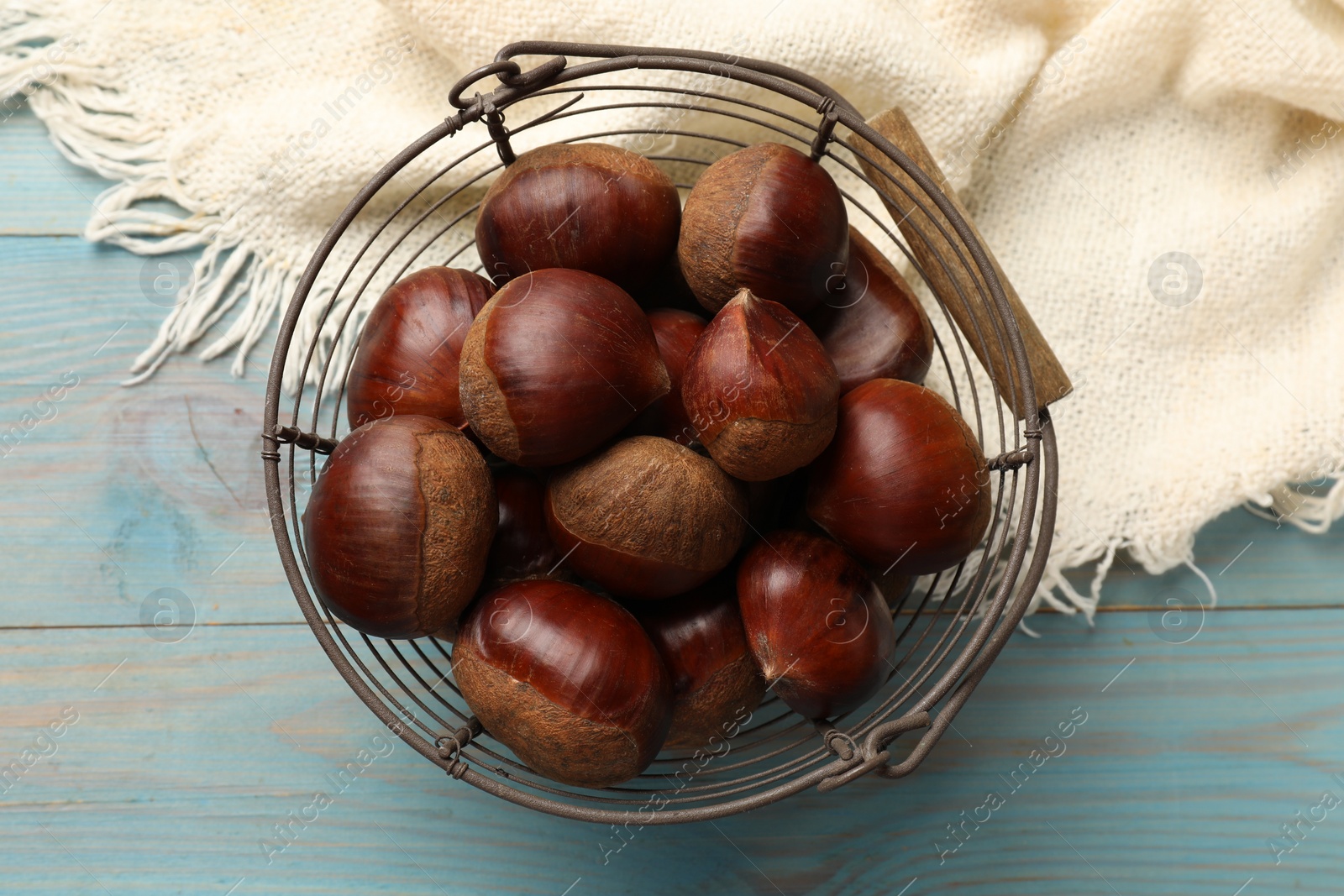 Photo of Roasted edible sweet chestnuts in metal basket on light blue wooden table, top view