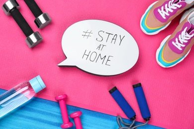 Photo of Sport equipment and speech bubble with hashtag STAY AT HOME on colorful yoga mats, flat lay. Message to promote self-isolation during COVID‑19 pandemic