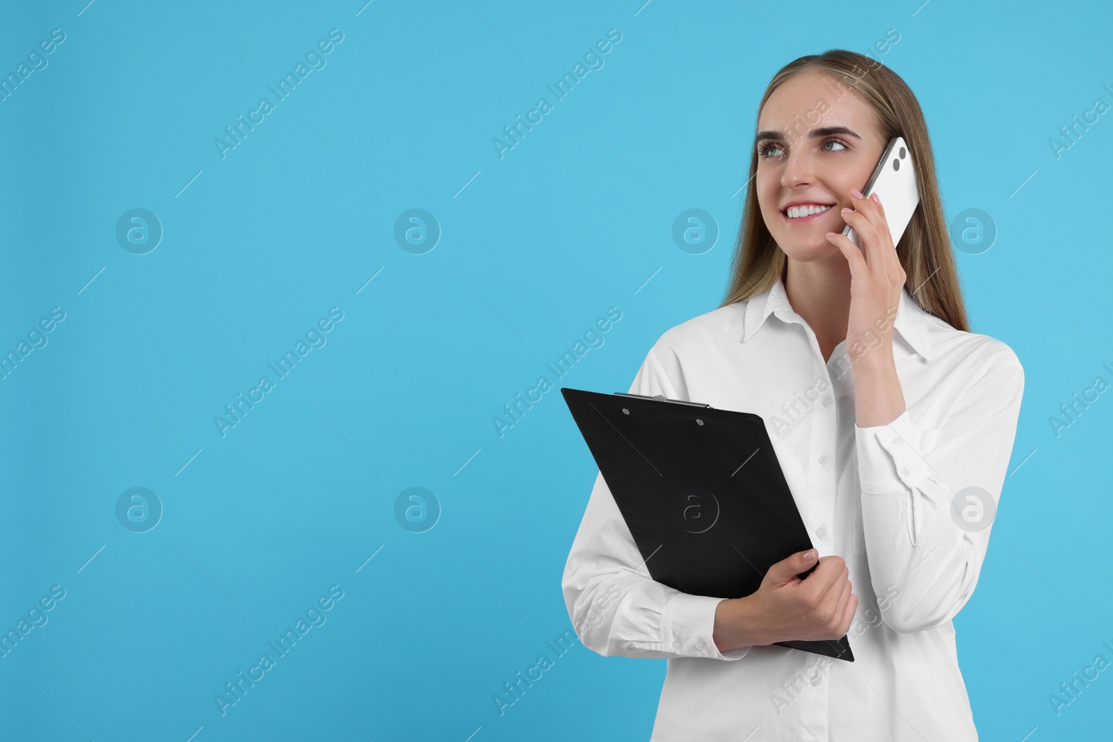 Photo of Happy young secretary with clipboard talking on smartphone against light blue background, space for text