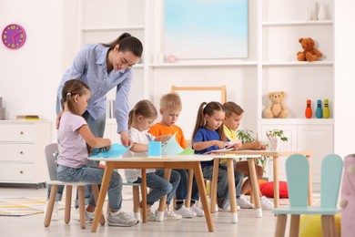 Photo of Nursery teacher with group of cute little children drawing and cutting paper at desks in kindergarten. Playtime activities