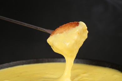 Dipping piece of bread into fondue pot with tasty melted cheese against dark gray background, closeup