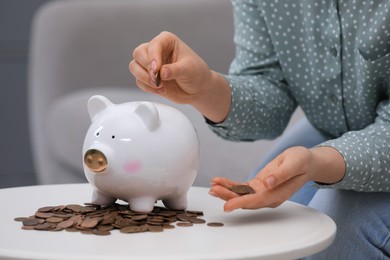 Young woman putting coin into piggy bank at table indoors, closeup