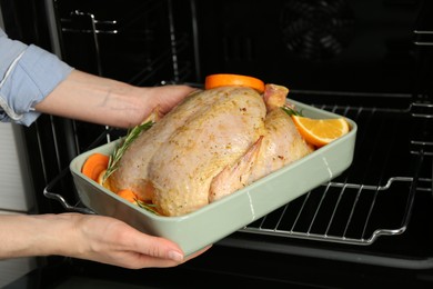 Photo of Woman putting pan with raw chicken and oranges into oven, closeup