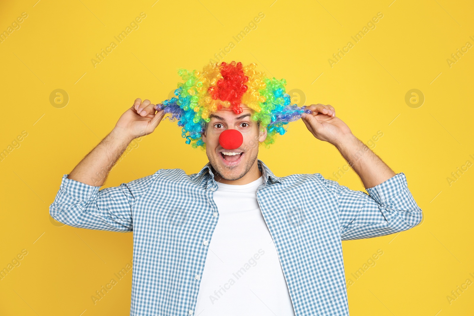 Photo of Funny man with clown nose and rainbow wig on yellow background. April fool's day