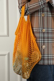 Woman holding mesh bag with different items near white wall, closeup. Conscious consumption