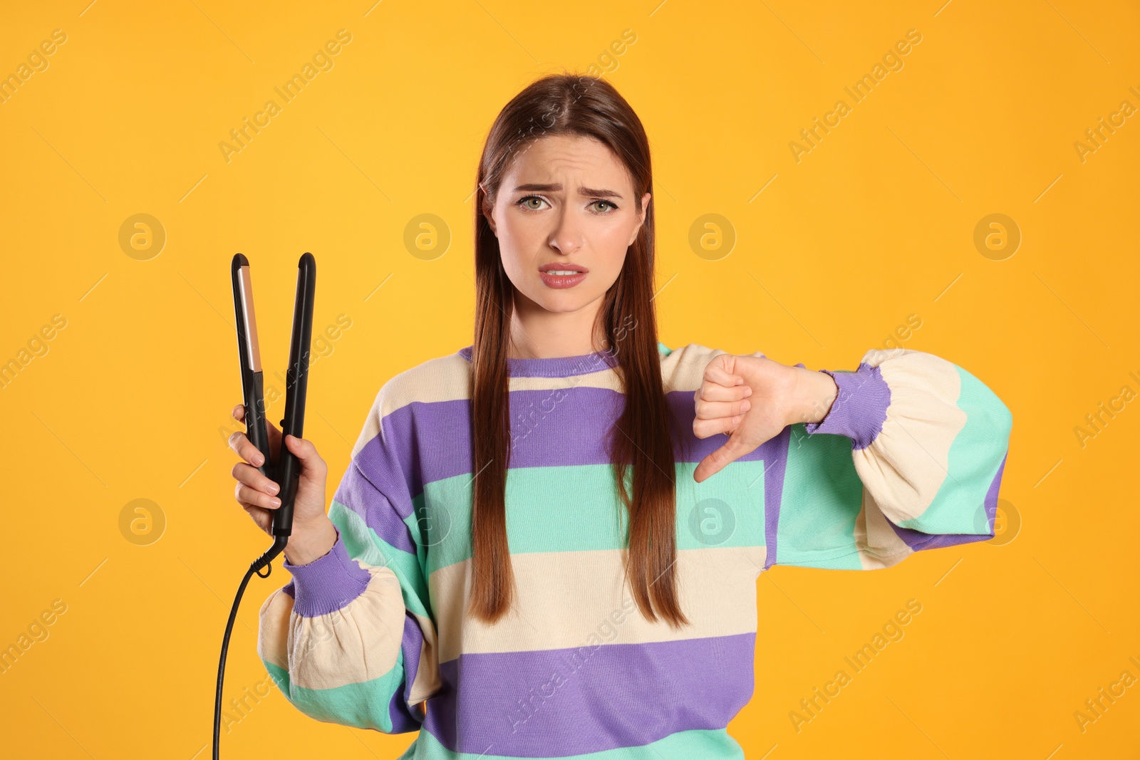 Photo of Upset young woman with flattening iron showing thumb down on yellow background. Hair damage
