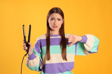 Upset young woman with flattening iron showing thumb down on yellow background. Hair damage